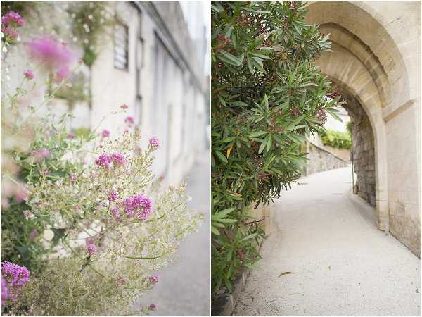 A diptych of two outdoor architectural detail shots with no people present. The left image is a close-up of wild pink and white flowering plants growing against a pale stone building wall, with shallow depth of field creating a soft, blurred background. The right image shows a stone archway passage with a curved vault leading to a pathway, flanked by a low stone wall and a large leafy shrub. Both shots highlight the venue's classic French stone architecture and grounds. Potential venue feature image.