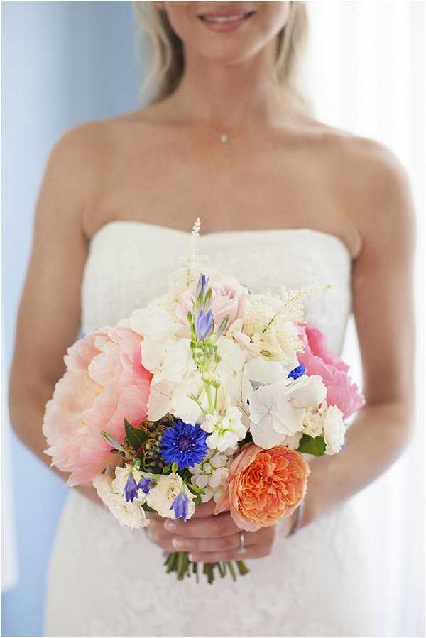 Close-up portrait of a bride from the shoulders down, holding a hand-tied bridal bouquet in front of her strapless white lace dress. The bouquet features blush pink peonies, a coral garden rose, white hydrangeas, bright cobalt blue cornflowers, pale blue agapanthus, cream stock flowers, small white astilbe sprays, and ivory sweet peas, creating a colorful summer palette. The bride wears a delicate pendant necklace and a ring is visible on her hand. The background is softly blurred in pale blue and white tones, consistent with an indoor getting-ready setting near a window.