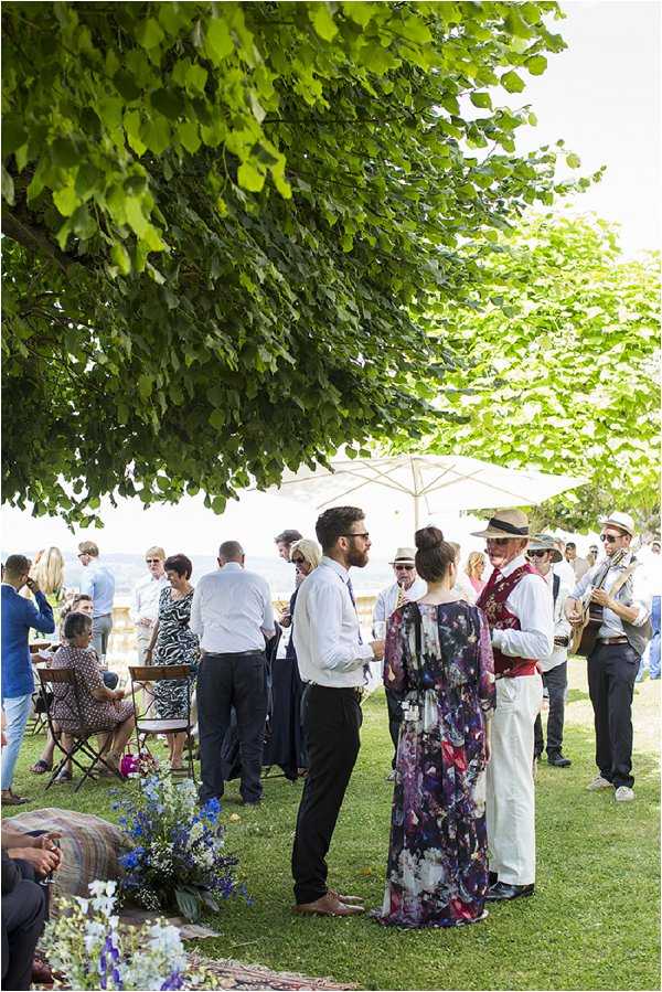 An outdoor cocktail hour taking place on a lawn with a water view in the background, with approximately 25–30 guests mingling in small groups. In the foreground, a man in a white shirt and dark trousers converses with a woman in a floral maxi dress, while nearby an older gentleman in a red vest and straw hat appears to be part of a live band, with another musician visible holding an instrument. Low floral arrangements featuring blue and white blooms, including what appear to be delphiniums and small white flowers, are placed along the ground in the foreground. White market umbrellas and bistro-style folding chairs are set up across the lawn. The overall styling is relaxed and informal, consistent with a garden party or boho-casual aesthetic. Wide shot capturing the full scene.