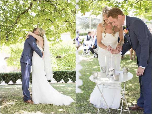 A two-image panel from an outdoor garden wedding ceremony. On the left, the bride and groom share a first kiss and embrace beneath a leafy tree; the bride wears a fitted, sleeveless white lace gown with a long train, and the groom wears a navy blue suit with brown shoes. On the right, the couple performs a unity sand ceremony at a small white bistro table, which holds two tall clear vases and several silver mercury-glass vessels; the bride wears a peach or coral flower accent in her hair, and the groom's boutonnière features a coral/peach bloom. White folding chairs with seated guests are visible in the background, indicating an outdoor lawn ceremony setting. Both images are close-to-medium portrait shots with bright, airy natural lighting and a classic, relaxed styling approach.