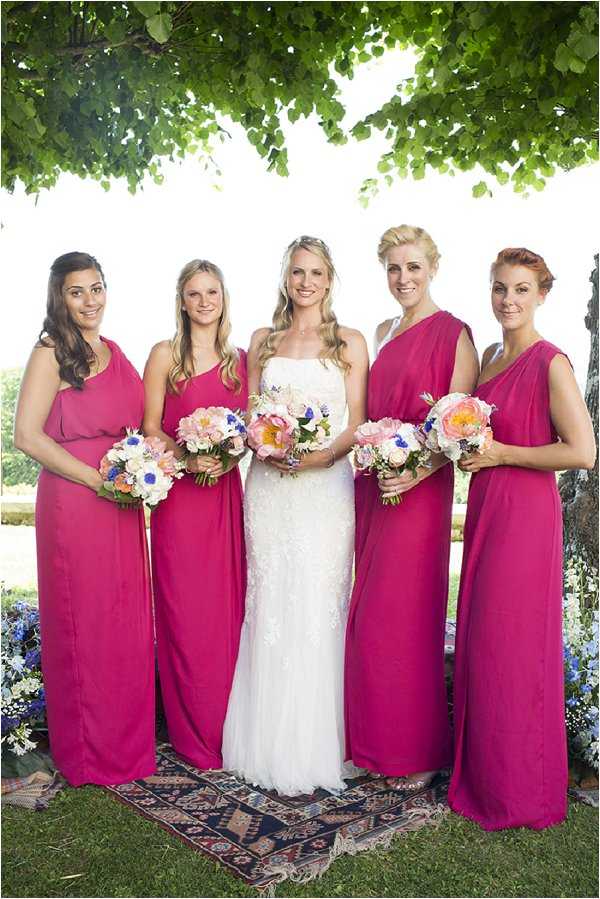 A bridal party portrait taken outdoors, featuring a bride and four bridesmaids standing together on a patterned rug laid on grass. The bride wears a fitted, strapless white lace gown and holds a bouquet of blush pink peonies, cobalt blue accents, and white flowers. The four bridesmaids wear floor-length fuchsia one-shoulder draped gowns and each carry matching bouquets of blush pink peonies, coral blooms, cobalt blue flowers, and white accents. The group is positioned under a large tree, with blue flowering shrubs visible in the background, suggesting a garden setting. The shot is a medium group portrait with bright, natural daylight.