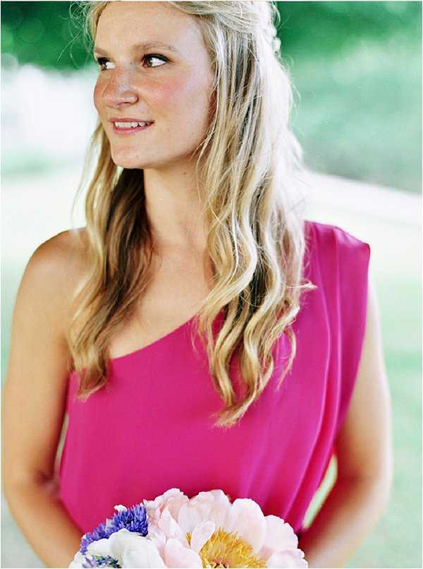 Close-up portrait of a bridesmaid with long, loosely wavy blonde hair worn down, dressed in a hot pink one-shoulder dress. She is holding a bouquet featuring blush pink peonies, purple cornflowers, and white blooms, and is looking slightly off-camera to one side. The image has a soft, film-quality look with a blurred green outdoor background, suggesting an outdoor setting. The overall styling is colorful and relaxed, with a summery, informal feel.