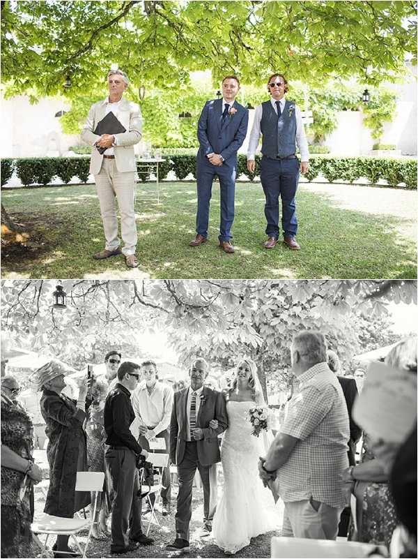A composite of two images from an outdoor garden wedding ceremony. The top image shows the groom in a navy blue suit with a pink boutonniere standing at the altar position alongside his best man in a navy blue vest and trousers, while the officiant in a beige linen suit holds a black folder; the setting is a manicured garden lawn shaded by a large tree, with white chair rows visible in the background. The bottom image is black and white and captures the bride being walked down the aisle by what appears to be her father, both facing the groom; the bride wears a fitted lace gown and carries a rounded bouquet, while seated and standing guests line the aisle on white folding chairs beneath the same large tree canopy. The ceremony has a relaxed, rustic-garden style with minimal visible decor beyond the natural setting.