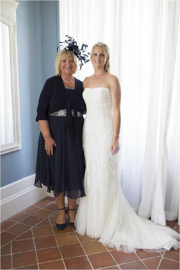 A portrait of two women posing together indoors in front of sheer white curtains, likely taken during the getting-ready portion of the wedding day. The bride wears a strapless, fitted ivory lace and tulle gown with a small train, while the older woman beside her — likely the mother of the bride — wears a knee-length navy chiffon dress with a navy feather and fascinator headpiece and a pearl necklace. The room features terracotta tile flooring and pale blue walls, with a white-framed mirror visible to the left. The image is a straightforward two-person portrait shot at mid-distance.