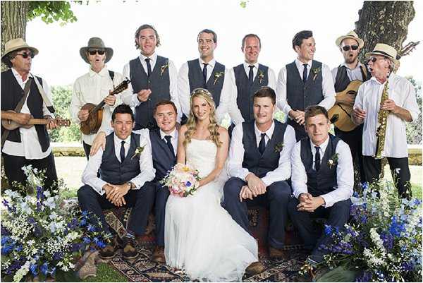 A group portrait taken outdoors featuring the bride and her groomsmen, plus what appears to be a live band, posed together on and around a patterned area rug in a garden setting. The bride wears a strapless white gown and holds a colorful bouquet of pink, coral, and white blooms with greenery, while the groomsmen are dressed in navy suits with charcoal waistcoats, white shirts, dark ties, and small floral buttonholes. Four musicians stand behind the group playing instruments including a ukulele, acoustic guitar, and saxophone, dressed casually in hats and white shirts. The foreground features clusters of cobalt blue delphiniums and white flowers arranged along the edge of the rug, adding a wildflower boho feel to the scene. Wide group shot with approximately 13 people visible.