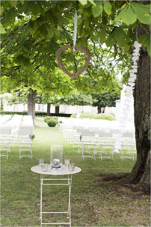 An outdoor wedding ceremony setup on a manicured lawn, with rows of white folding chairs arranged in an aisle formation. In the foreground, a small white bistro table holds glass votive candle holders and a hurricane lantern centerpiece. A woven twig heart wreath is suspended by a white ribbon from a large tree branch overhead, serving as a decorative focal point. The styling is classic and understated, with an all-white furniture palette and natural wicker accents, set against a formal garden backdrop with trimmed hedges and a white building visible in the distance. Wide shot taken from beneath the tree canopy looking out over the ceremony space. Potential venue feature image.
