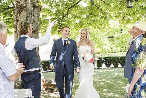 The bride and groom walk back up the aisle following an outdoor ceremony, greeted with celebratory cheers and confetti from guests on either side. The ceremony takes place in a garden setting beneath a large leafy tree, with trimmed hedges and hanging lanterns visible in the background. The bride wears a fitted strapless white gown and carries a bouquet of peach, coral, and pink blooms, while the groom wears a navy blue suit with a peach boutonniere. Approximately four to five guests are visible in the foreground, dressed in a mix of casual and colorful attire, with the group captured in a mid-shot candid composition.
