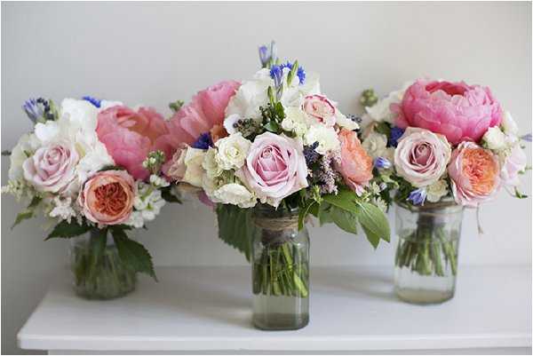 A flat-lay-style detail shot of three bridal bouquets displayed in glass mason jars on a white surface against a plain light wall. The bouquets feature a mix of large hot pink peonies, mauve and blush roses, peach garden roses, white lisianthus, blue delphinium, and dark berry-toned filler flowers with green foliage. The central bouquet is the largest and most elaborate, wrapped with twine at the neck of the jar, suggesting it is the bridal bouquet, while the two flanking arrangements are slightly smaller and serve as bridesmaid bouquets. The overall floral palette is a soft garden-style mix of pink, peach, mauve, white, and blue, consistent with a romantic, garden-inspired wedding aesthetic.