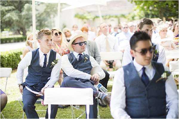 Guests are seated outdoors during a wedding ceremony, watching proceedings from white folding chairs arranged in rows. The setting appears to be a garden or grounds under bright sunlight with a white canopy or tent visible in the background. In the foreground, three young men wearing matching navy waistcoats, white dress shirts, and navy ties with small floral buttonnieres are visible — likely groomsmen — with two wearing sunglasses including one in a straw fedora hat. Behind them, a larger crowd of approximately 30 or more guests is visible, dressed in summer attire. The shot is a medium wide-angle taken from a low angle, looking back toward the seated guests rather than toward the altar.