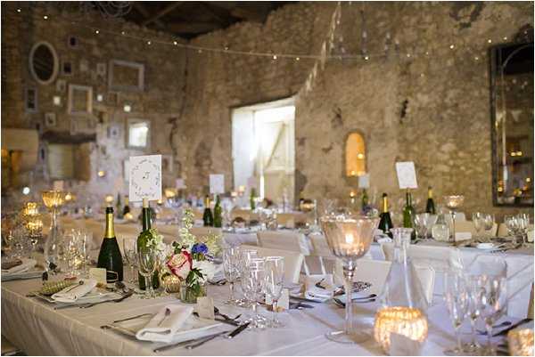 A wide interior shot of a wedding reception set up inside a rustic stone-walled venue, likely a converted barn or historic French building. Long rectangular tables are dressed in white linen and set with crystal glassware, white folded napkins, silver cutlery, and gold mercury glass votive candles, with champagne bottles placed along the tables as decor. Centerpieces feature small arrangements of pink, red, and blue mixed flowers in low vases. The rough stone walls are decorated with an eclectic collection of white-painted picture frames and small artworks, and warm fairy lights are strung across the ceiling alongside what appears to be white bunting or paper garland. The overall decor palette is white and gold with warm candlelight, giving the space a rustic yet refined feel. Potential venue feature image.