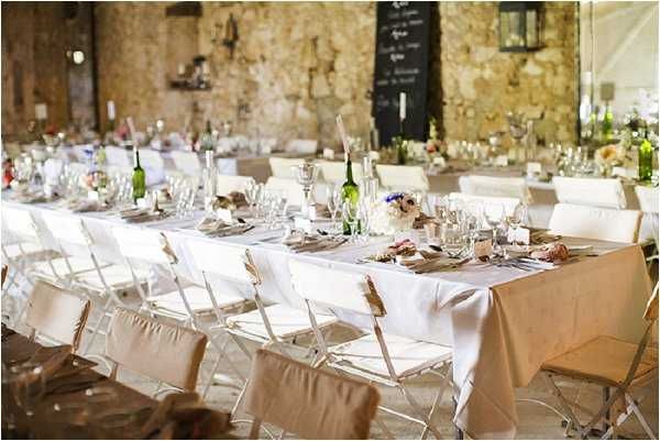 A wide shot of a wedding reception dining setup inside a rustic stone-walled venue, likely a converted barn or mas. Multiple long rectangular tables are dressed in white tablecloths with taupe/sand linen runners, set with glassware, silver cutlery, and low floral centerpieces featuring white blooms and touches of blush and blue. White folding chairs with taupe fabric seat cushions are arranged along both sides of the tables. Green wine bottles are used as decorative elements among the table settings, and a chalkboard menu sign is visible on the back wall. The overall decor palette is neutral — white, sand, and taupe — with a rustic-chic styling theme. A lantern-style light fixture is mounted on the stone wall in the background.