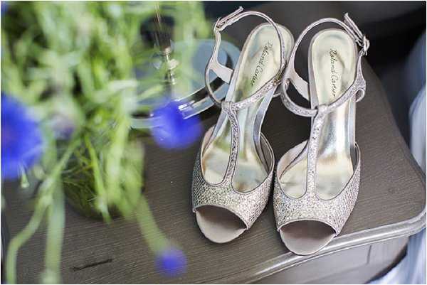 A close-up detail shot of a pair of silver glittery open-toe T-strap heeled sandals, labeled René Caovilla, placed on a dark wooden surface. In the soft-focus foreground, vivid cobalt blue flowers — likely cornflowers or nigella — and green foliage are visible, along with a blurred metallic candleholder in the background. The overall styling suggests a blue and silver decor palette.