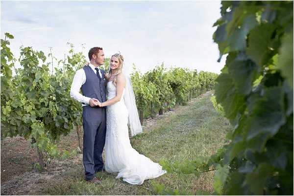 A couple portrait taken outdoors in the rows of a vineyard during what appears to be late afternoon light. The bride wears a fitted ivory lace gown with a cathedral-length veil and a floral hair accessory, while the groom is dressed in a grey waistcoat, white dress shirt, dark trousers, and a boutonnière. The two stand close together, with the bride leaning into the groom as they look at each other. The shot is a medium-wide portrait framed by vineyard foliage in the foreground, with rows of vines receding into the background.