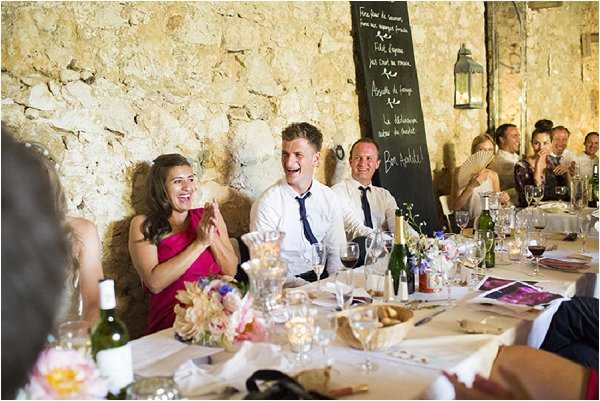A wedding reception dinner is underway inside a rustic stone-walled venue, likely a French chateau or barn, with guests seated at a long rectangular table set with wine bottles, glassware, candles, and small floral centerpieces featuring pink peonies and mixed blooms. A man in a white shirt and navy tie at the center of the frame is laughing openly, while the woman beside him in a fuchsia dress claps with delight, suggesting a speech or toast is being delivered. Approximately 10-15 guests are visible, with a large blackboard menu written in French chalked with the words 'Bon Appétit' propped against the back wall, and a metal lantern hanging nearby. The shot is a medium-wide candid taken from across the table, capturing the relaxed and jovial atmosphere of the meal.
