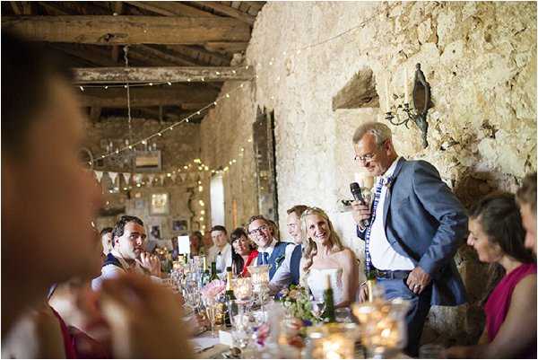 A wedding speech is taking place during a reception dinner inside a rustic stone barn venue. A man in a grey suit holding a microphone stands at the end of a long banquet table delivering his speech, while the bride in a white strapless dress and the groom in a blue waistcoat sit among guests laughing and listening. The table is dressed with candlelight from gold votives, glassware, bottles, and what appear to be loose floral centerpieces in pink and green tones. The venue features exposed stone walls, heavy wooden ceiling beams, and strings of warm fairy lights along with colorful bunting banners hanging overhead, creating a rustic, informal atmosphere. The shot is taken from a slightly elevated wide angle, with a blurred guest in the foreground framing the scene.