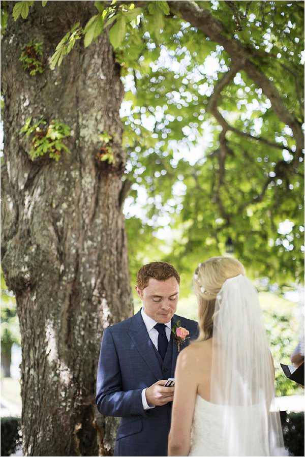 An outdoor wedding ceremony takes place beneath a large tree, with the groom reading his vows from a small card or booklet. The groom wears a navy blue three-piece suit with a dark navy tie and a coral/peach rose boutonniere. The bride stands with her back to the camera, wearing a white lace gown and a long cathedral-length veil with a pearl or crystal hair accessory at the crown. A third person, likely the officiant, is partially visible at the far right edge of the frame. The composition is a medium portrait shot with the tree trunk dominating the left side and bokeh foliage filling the background.