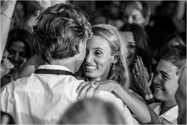 Black-and-white close-up portrait of a bride and groom embracing on the dance floor, surrounded by a crowd of guests during what appears to be the first dance or open dancing at a reception. The bride is smiling broadly and wearing a delicate headband tiara and drop earrings, with her arms around the groom who has his back to the camera. A young boy and several other guests are visible in the background, also smiling and reacting to the moment. The image has strong contrast with bright highlights on the couple and softer tones in the crowd behind them.