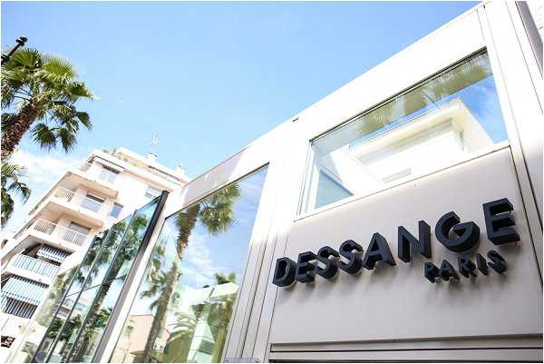 This image shows the exterior facade of a Dessange Paris salon, a hair and beauty brand, photographed from a low angle in bright daylight. The building features a modern white and glass storefront with bold black three-dimensional lettering reading 'DESSANGE PARIS.' This does not appear to be a wedding photograph; it is an exterior commercial building shot with no people, wedding attire, decor, or ceremony elements visible.