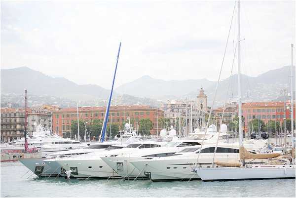 Wide shot of a marina in what appears to be Nice, France, showing multiple large white motor yachts and sailing vessels moored at dock, with the city's distinctive ochre and terracotta-colored buildings visible in the background along with tree-lined streets and mountain ranges in the distance. The image does not appear to contain any wedding subjects, people, or wedding-related activity — no couple, guests, decor, or ceremony elements are visible. The shot functions as a location or venue context image, likely used to establish the South of France coastal setting for a wedding feature. Potential venue feature image.