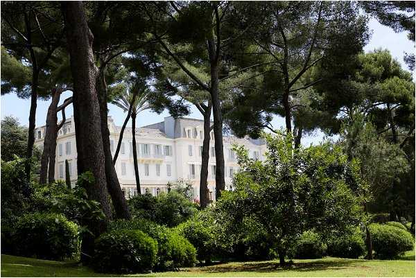 Wide exterior shot of a large white French chateau or villa viewed through mature pine and deciduous trees, with manicured shrubs and a green lawn in the foreground. The building features classic French architecture with tall white-framed windows, grey roofing, and pale yellow facade accents. No people or wedding-specific elements are visible in this image. Potential venue feature image.