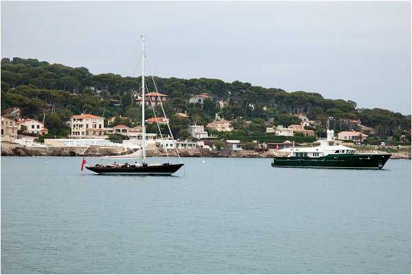 This image does not appear to be a wedding photograph. It shows a coastal seascape with two boats — a black sailing yacht and a dark green motor yacht — anchored in calm blue-green water, with a shoreline village visible in the background featuring residential buildings and dense tree coverage. There are no people, wedding attire, decor, or any wedding-related elements visible in this image.