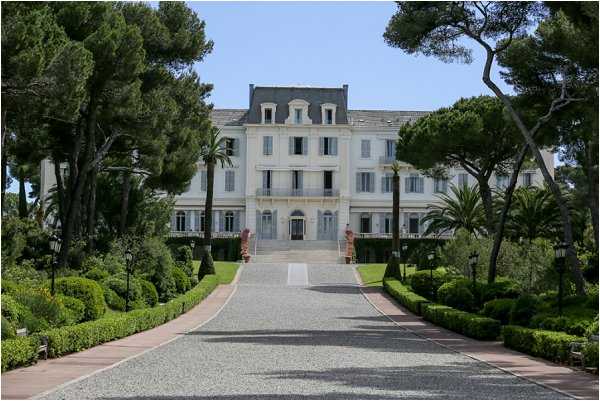 A wide exterior shot of a large white French-style villa or chateau, photographed straight-on from the end of a long gravel driveway. The building is a multi-story pale white structure with a mansard roof, shuttered windows, wrought-iron balconies, and a grand central entrance approached by a broad stone staircase. The grounds are formally landscaped with neatly trimmed box hedges, manicured lawn borders, lamp posts lining the driveway, and mature Mediterranean pine and palm trees framing the facade. No people are visible in the image. Potential venue feature image.