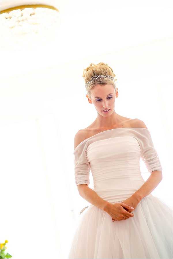 A bridal portrait of a single bride standing indoors in a bright, window-lit interior space with a chandelier partially visible in the upper left corner. The bride wears an off-the-shoulder white ball gown with a ruched bodice, three-quarter length sleeves, and a full tulle skirt. Her blonde hair is styled in an updo adorned with a delicate crystal and pearl headband tiara. She is looking downward with her hands clasped at her waist. The image is slightly overexposed, giving it a very bright, airy feel. Medium portrait shot from approximately the hips up.