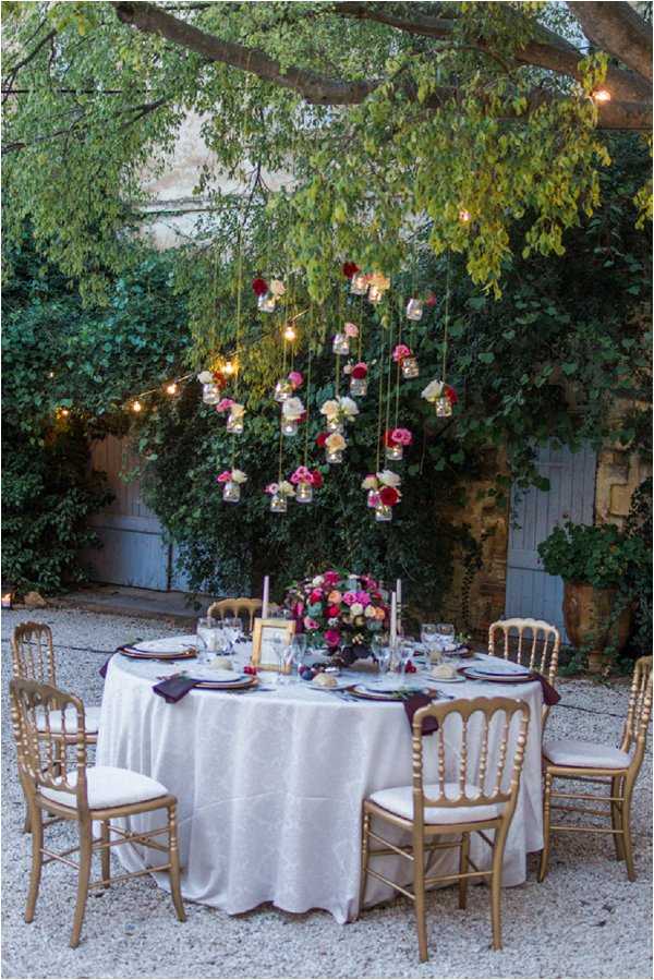 An outdoor wedding reception table styled in a courtyard or garden setting, photographed at dusk as a wide detail shot. The round table is dressed in a white damask linen with deep burgundy/plum napkins, gold charger plates, glassware, and a lush centerpiece of hot pink, deep red, blush, and cream roses. Six gold Chiavari chairs with cream cushions surround the table. Above the table, small glass jars holding individual blooms in matching shades of fuchsia, pink, and white are suspended at varying heights from a tree branch, interspersed with warm Edison-style globe lights and string lights. Lavender taper candles are placed on the table. The overall decor palette is jewel-toned with gold accents, leaning toward a romantic garden style.