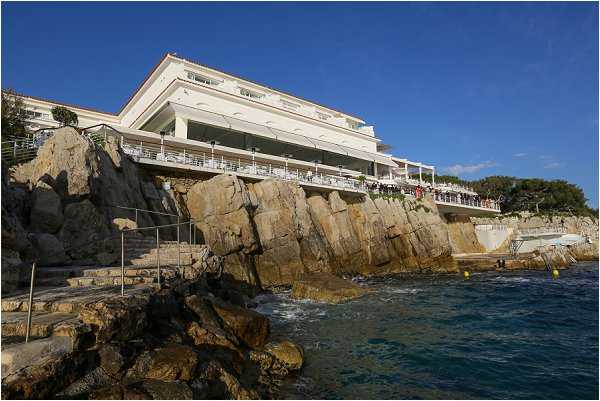Wide-angle exterior shot of a large white multi-story hotel or venue building perched directly on rocky coastal cliffs above the sea. A group of people, likely wedding guests, are visible gathered on one of the venue's terraces or balconies along the seafront facade. The building features multiple levels of white-railed terraces overlooking the water. The shot is taken from water level or a low coastal vantage point, emphasizing the dramatic positioning of the venue on the rocks above the Mediterranean. Potential venue feature image.