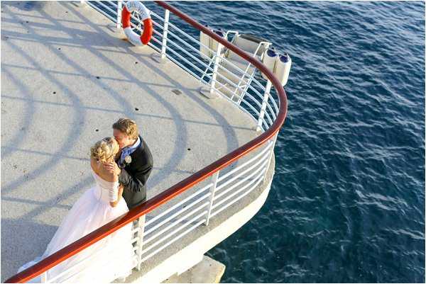 A couple shares a kiss during wedding portraits on the curved deck of a boat or yacht, photographed from an elevated angle looking down. The bride wears a white strapless ball gown and has blonde hair styled up, while the groom is dressed in a dark suit. A red curved railing runs along the white deck, and a life preserver ring is visible in the background. The deep blue water is visible beside the vessel. Wide shot taken from above, giving a bird's-eye perspective of the couple positioned at the bow of the deck.