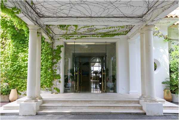 Wide shot of a venue entrance featuring a white colonnaded portico with classical white columns and marble steps leading to glass entry doors. The pergola overhead is covered in bare branching vines, while the surrounding walls are heavily draped in lush green ivy. Through the glass doors, a glimpse of an interior space with warm lighting and arched architectural details is visible. No people are present in the image. Potential venue feature image.