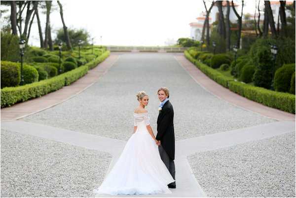 A couple portrait taken outdoors on a wide gravel driveway flanked by symmetrically trimmed hedges and topiary, leading toward a formal garden and a villa building visible in the background. The bride wears an off-the-shoulder white ballgown with a full tulle skirt and has her hair pinned up, while the groom wears a classic black tuxedo with a white shirt. Both are holding hands and have turned to look back toward the camera, posed mid-walk along the central axis of the driveway. The composition is a wide shot emphasizing the formal landscaped grounds, with the couple positioned in the lower center of the frame. Potential venue feature image.