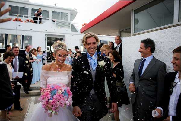 The bride and groom walk through a confetti shower immediately following their ceremony, surrounded by approximately 15-20 smiling guests on either side forming a receiving line. The setting is outdoors on a dock or marina, with a large white and red ferry or yacht vessel visible directly behind the couple. The bride wears an off-the-shoulder white gown with a sheer wrap, her hair styled up with a decorative piece, and carries a large cascading bouquet of pink and blue hydrangeas and flowers. The groom wears a dark navy suit with a light blue tie and a white boutonniere. Bridesmaids in pale blue dresses are visible in the background. The shot is a wide portrait-style image capturing the couple mid-stride with confetti filling the air around them.