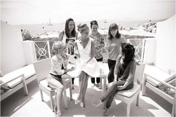 A black-and-white getting-ready moment showing a bride seated at the center of a group of five women on an outdoor terrace overlooking the sea. The bride wears a short, full-skirted lace or embroidered dress with a V-neckline and a decorative headpiece, while the other women are dressed casually in jeans, shorts, and patterned tops. The terrace features white wicker and rattan chairs with cushions, and the background reveals a coastal landscape with rocky outcroppings and sailboats on the water. The image is a wide group shot with high-key, bright tones and soft contrast typical of natural daylight photography.