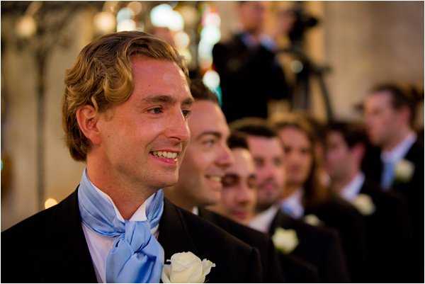 The groom stands at the altar inside what appears to be a church or chapel, smiling as he watches the bride approach. He is wearing a dark suit jacket with a pale blue cravat and a white rose boutonniere. Behind him, a row of at least five groomsmen are visible, all dressed in matching dark suits with pale blue ties and white rose boutonnieres. The interior setting shows warm ambient lighting typical of a traditional church ceremony. Close-up portrait shot focused on the groom with the groomsmen receding in the background.