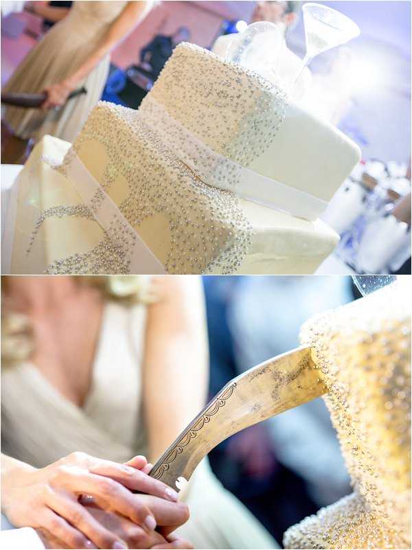 A two-image collage capturing the wedding cake cutting moment at an indoor reception. The top image shows a close-up detail shot of a three-tiered square white fondant wedding cake decorated with cascading silver and pearl beaded embellishments and ivory satin ribbon bands between each tier, with a glass topper visible and guests in the background. The bottom image is a close-up of the couple's hands holding an ornate engraved silver cake cutting knife as it touches the edge of the beaded cake, with the bride visible in a sleeveless ivory gown with a cowl neckline. The overall decor palette is white, ivory, and silver with a modern classic styling.