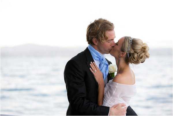 A close-up portrait of a bride and groom sharing a kiss outdoors with a wide expanse of open water visible in the soft-focus background. The groom wears a dark suit jacket with a light blue cravat-style tie and a white rose boutonniere. The bride wears an off-the-shoulder white dress and has her blonde hair styled in an updo accessorized with a crystal or rhinestone tiara headband; her engagement ring is visible as she rests her hand on the groom's chest. The image is slightly overexposed, creating a bright, airy feel with pale tones throughout.