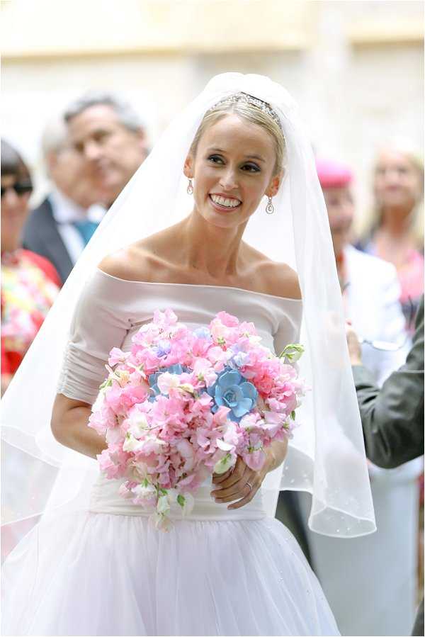 Close-up portrait of a smiling bride walking, likely during a ceremony procession, with a crowd of guests visible in the soft-focus background outdoors. She wears an off-the-shoulder white gown with a fitted bodice and a full tulle skirt, paired with a cathedral-length veil and a delicate diamond tiara, and drop earrings. Her bouquet is a large, rounded arrangement of pink sweet peas and blue succulents. The styling is classic and formal in feel, with the floral palette combining soft pink and powder blue tones.