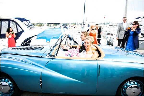 A bride and groom are seated in a vintage powder-blue convertible sports car, likely from the 1950s, driving through a marina setting surrounded by moored boats and yachts. The bride, wearing a strapless white dress and holding a pink and white bouquet, is laughing, while the groom sits beside her. Several guests line the route in the background, including a woman in a red dress, a woman in an orange dress, a man in a grey suit, and a woman in a teal dress, some holding drinks and glasses. The wide shot captures a lively send-off moment with confetti visible in the air around the car.