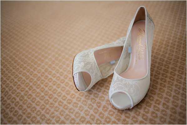 A close-up detail shot of a pair of bridal heels placed on a textured beige patterned surface. The shoes are ivory lace peep-toe pumps with a pink insole lining and a stiletto heel, featuring delicate floral lace overlay on the upper. A small blue ribbon detail is visible inside one shoe, likely a nod to the 'something blue' tradition. The composition is a flat lay-style detail shot with a shallow depth of field, keeping the foreground shoe in sharp focus.