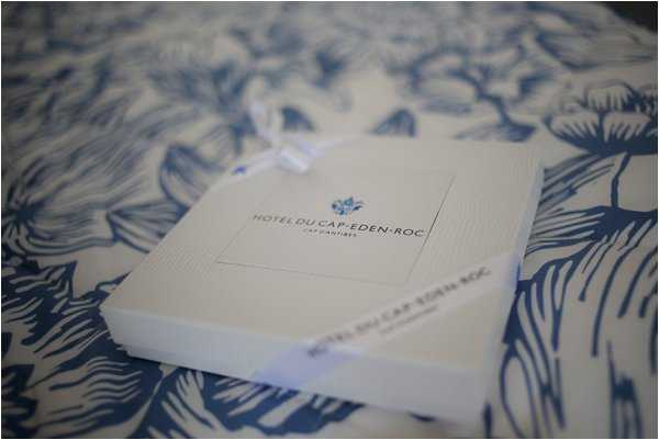 A close-up detail shot of two white gift or stationery boxes from the Hôtel du Cap-Eden-Roc, Cap d'Antibes, tied with a white satin ribbon bow, resting on a blue and white floral-patterned fabric or surface. The boxes feature the hotel's logo and name printed in blue text. The composition is a shallow depth-of-field detail shot with soft, slightly dim lighting, giving the background a blurred effect.
