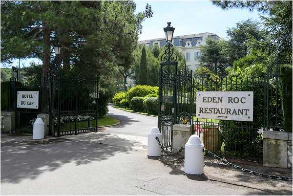Wide shot of the entrance gates to the Hôtel du Cap and Eden Roc Restaurant, showing ornate black wrought-iron gates flanked by stone pillars with lantern-style lamp posts and white bollards. A paved driveway leads through the gates toward a large white multi-story building visible through the tree-lined grounds. No people or wedding activity are present in this image. Potential venue feature image.