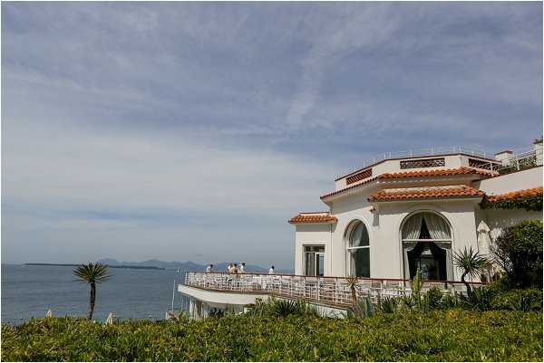 Wide exterior shot of a white Mediterranean-style villa with terracotta roof tiles, featuring a curved terrace overlooking the sea. A small group of people dressed in white or light-colored attire are gathered on the terrace, likely during a cocktail hour or pre-ceremony gathering. The building has arched windows and a rooftop balcony with decorative railings, set directly on the coastline with a panoramic sea view and distant islands visible on the horizon. Potential venue feature image.