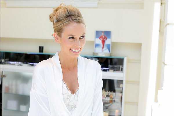 A bride is photographed during the getting-ready stage, wearing a white satin robe with lace trim underneath. Her blonde hair is styled in an upswept bun. She is indoors in what appears to be a hair and beauty salon or dressing room, with shelving units holding beauty products and equipment visible in the background. The shot is a close-up portrait with soft, bright natural lighting giving the image a light, airy feel.