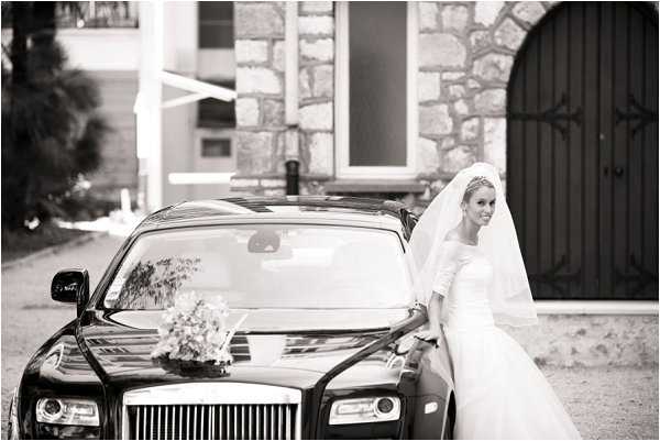A black-and-white portrait of a bride standing beside a Rolls-Royce luxury car, leaning slightly against the vehicle and looking toward the camera. The bride wears a long-sleeved white gown with a long cathedral veil and a decorative headpiece, and a bridal bouquet rests on the hood of the car. The setting appears to be the entrance of a stone-facade building with large dark wooden doors visible in the background. The image is a medium-wide shot with soft contrast and good tonal range throughout.