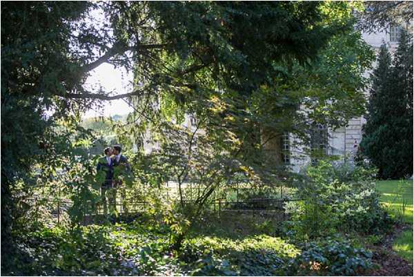 A couple shares a kiss during outdoor wedding portraits in the grounds of what appears to be a French chateau or manor house, visible in the background with pale stone facade and tall windows. The pair are partially obscured by dense garden foliage and stand near a wrought-iron gate or fence, framed by large mature trees. The groom wears a dark navy suit and the bride wears a fitted dark outfit, though detail is limited due to distance. This is a wide environmental shot with the couple as a small focal point within the lush garden landscape, taken in bright dappled natural light. Potential venue feature image.