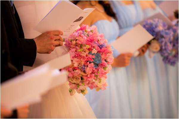 A close-up detail shot taken during a wedding ceremony, showing the bride and bridesmaids standing in a row holding white ceremony programs. The bride carries a full, rounded bouquet of hot pink and blush sweet peas, pink roses, and blue hydrangea accents. The bridesmaids are dressed in pale powder blue strapless gowns and hold smaller bouquets of purple and blue hydrangeas. The composition focuses on the mid-section of the group, cutting off faces, with a shallow depth of field that softly blurs the background figures.