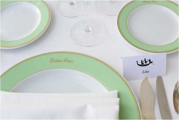 Close-up detail shot of a formal wedding reception table setting featuring branded Eden Roc crockery — white plates with a mint green and gold rim — arranged on a white linen tablecloth. A folded white napkin sits on the main charger plate, alongside silver cutlery and clear crystal glassware. A white place card printed with a gondola illustration and the guest name 'Lita' is propped against the setting, suggesting a Venetian or nautical theme. The overall decor palette is mint green, gold, and white.