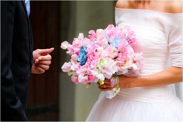 A close-up portrait shot of a bride and groom standing together, cropped at the torso with neither face visible. The bride wears an off-the-shoulder white dress with ruched sleeves and holds a rounded bouquet composed of pink and pale pink sweet peas, blue hydrangeas, and soft blush garden roses. The groom, wearing a dark navy suit, extends one hand toward the bride. The background is softly blurred with a dark door or wall visible behind them.