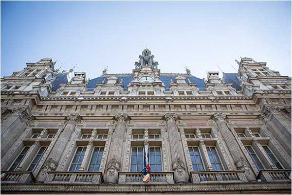 Wide-angle upward-facing shot of a grand French Beaux-Arts or Second Empire-style civic building, likely a mairie (town hall), featuring ornate stone facade with carved detailing, tall arched windows, a mansard roof with dormer windows, a central clock, and a French tricolor flag displayed on the balcony. No people or wedding party are visible in the frame. The image appears to be taken at the exterior of a civil ceremony venue. Potential venue feature image.